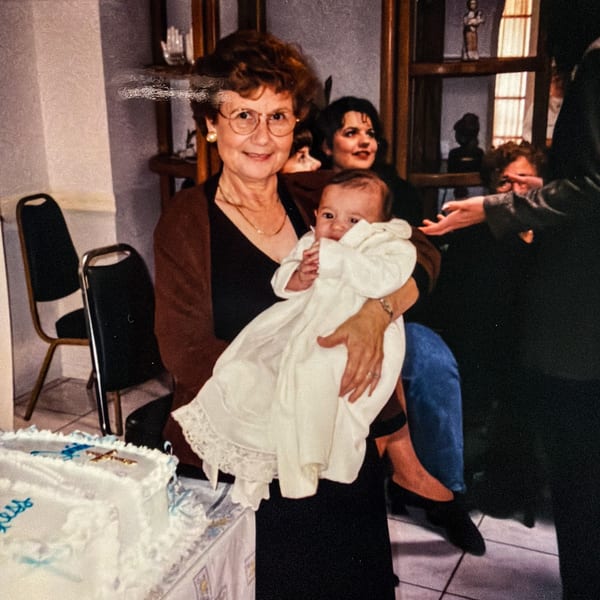 A woman in glasses holds a baby in a white gown, with a cake nearby.