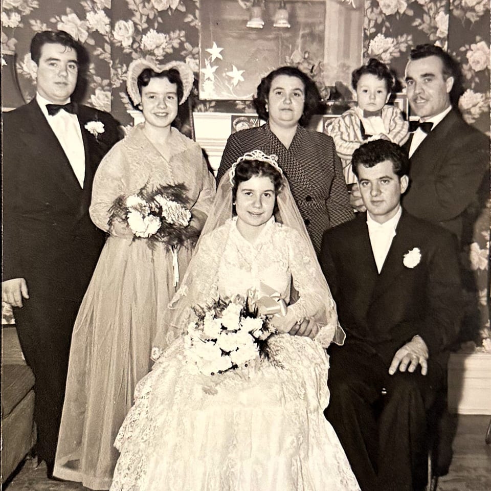A vintage wedding photo shows a bridal party of seven, including a baby, posing indoors.