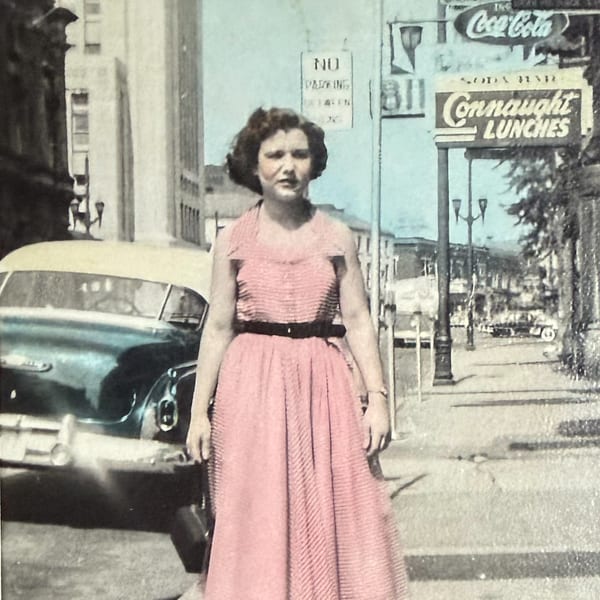 A woman in a pink dress stands on a street with a vintage car and "Connaught Lunches" sign.