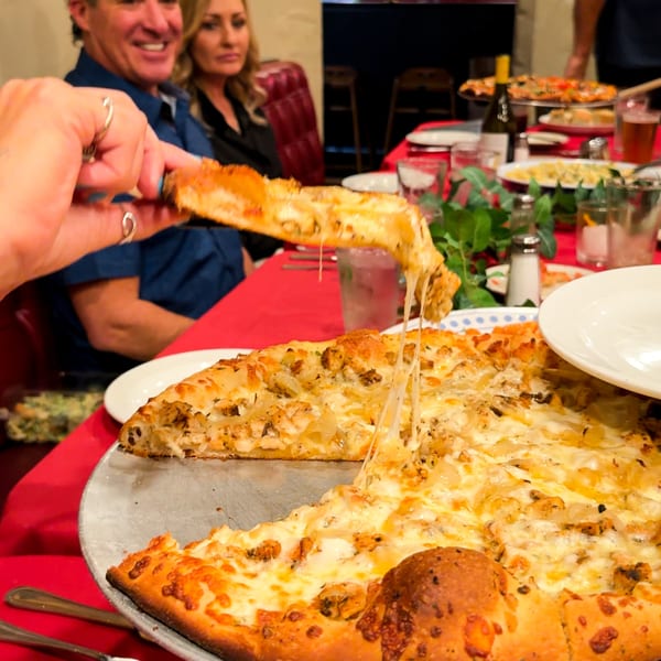 Hand pulling a cheesy slice of chicken and onion pizza from a large pie on a red tablecloth.