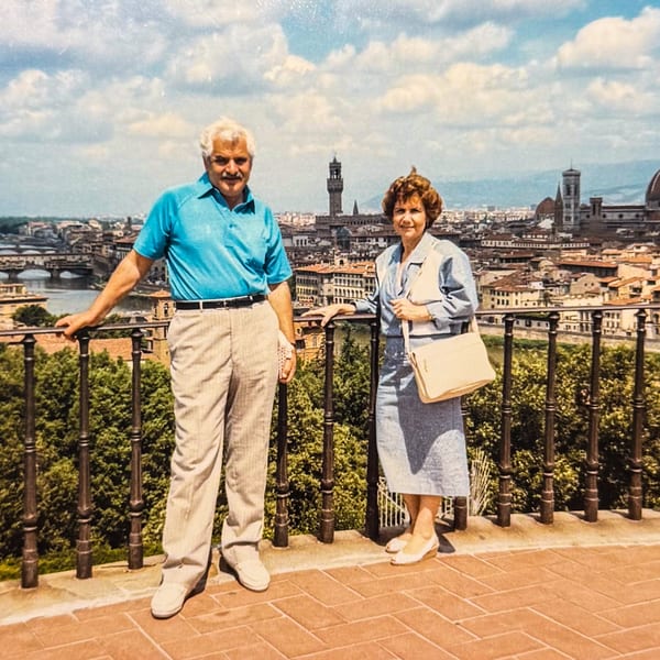 A couple stands on a balcony overlooking a European city with a river and historic buildings.