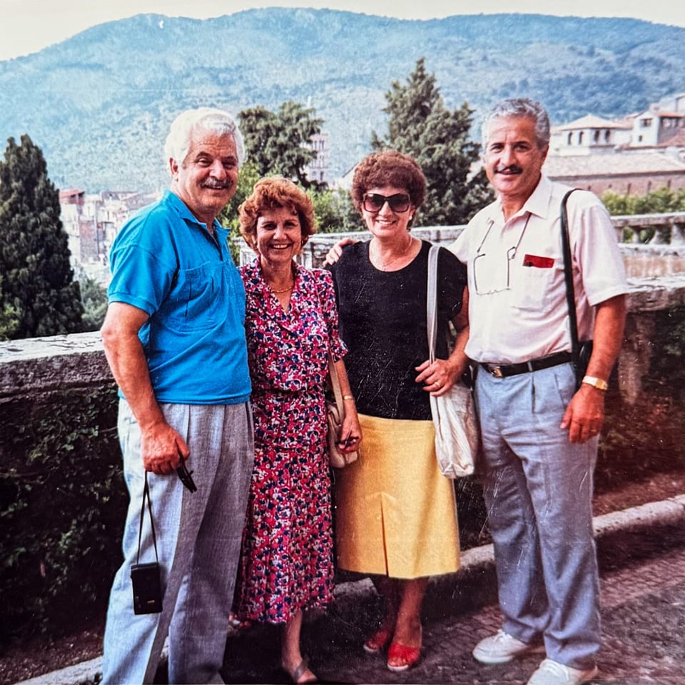 Two couples smile at the camera from a stone balcony overlooking a mountainous town.