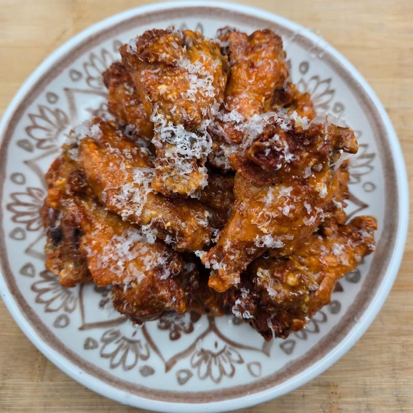 A plate of crispy, glazed chicken wings topped with shredded white cheese on a patterned plate.