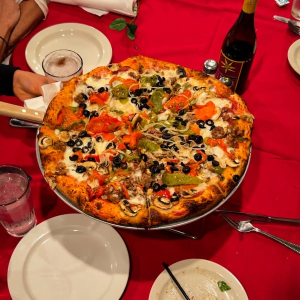 A large, sliced pizza with various toppings sits on a red tablecloth with drinks and plates.