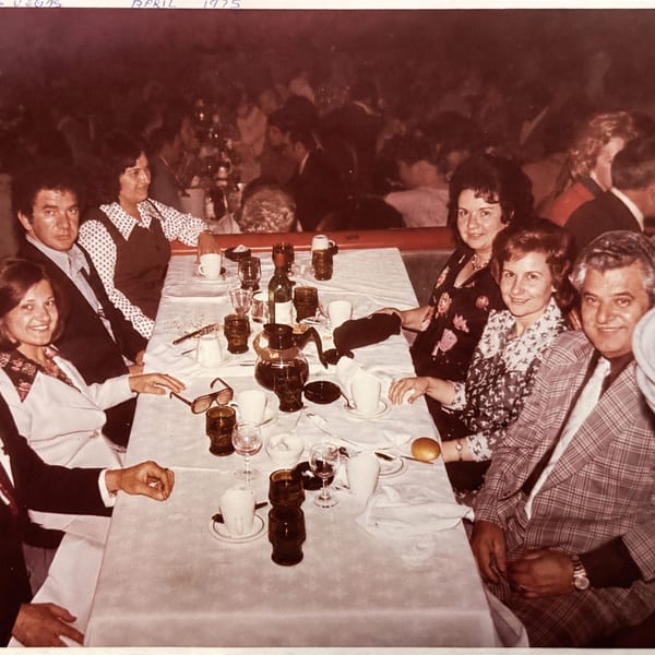A group of six people seated around a white-clothed table with drinks and dinnerware.