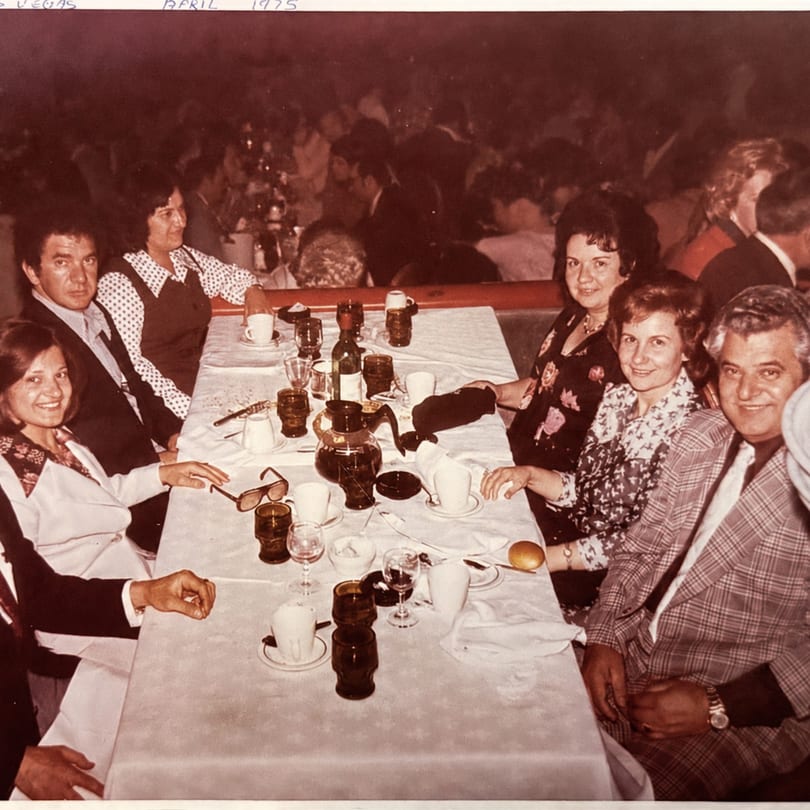 A group of six people seated around a white-clothed table with drinks and dinnerware.