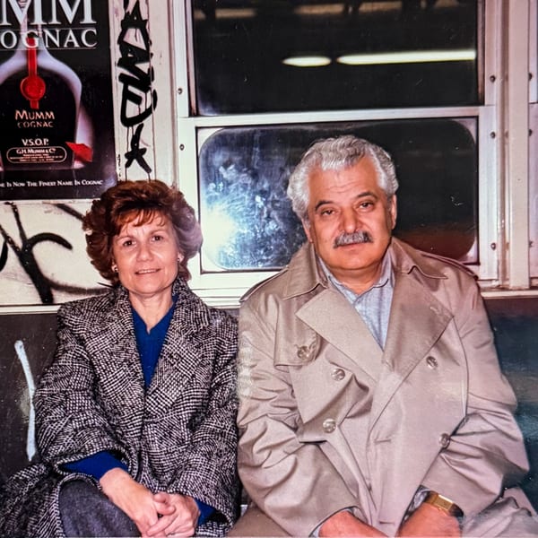 A smiling couple, a woman in a patterned coat and a man in a trench coat, sitting on a subway.