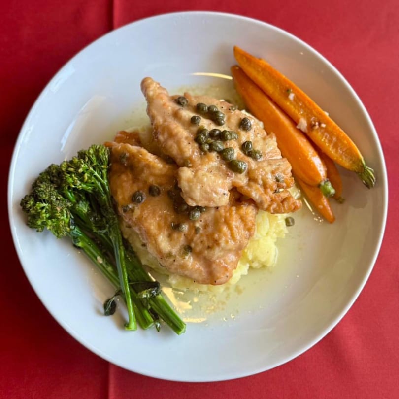 Chicken piccata with capers, mashed potatoes, broccolini, and glazed carrots on a white plate.