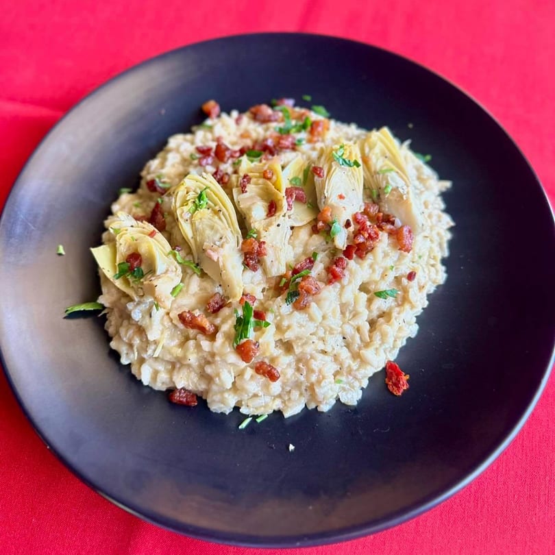 Creamy risotto topped with artichoke hearts, crispy bacon bits, and fresh parsley on a black plate.