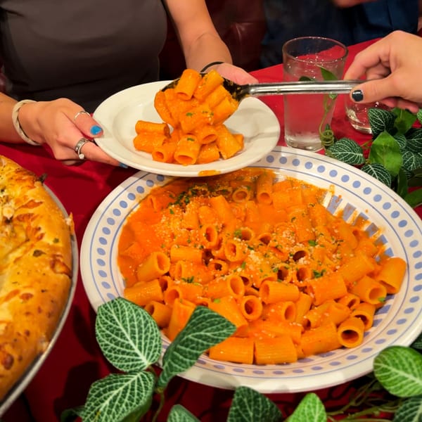 Hands serving rigatoni pasta with red sauce and parmesan cheese from a large bowl onto a plate.