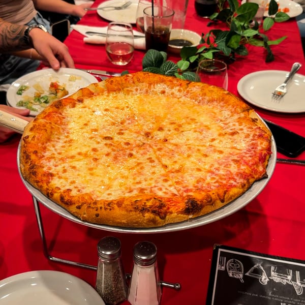 A large cheese pizza on a stand, with diners and drinks around a red-clothed table.