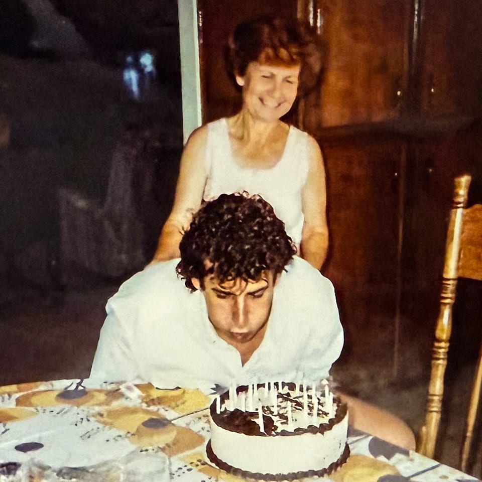 A man blows out candles on a birthday cake while a woman smiles behind him.