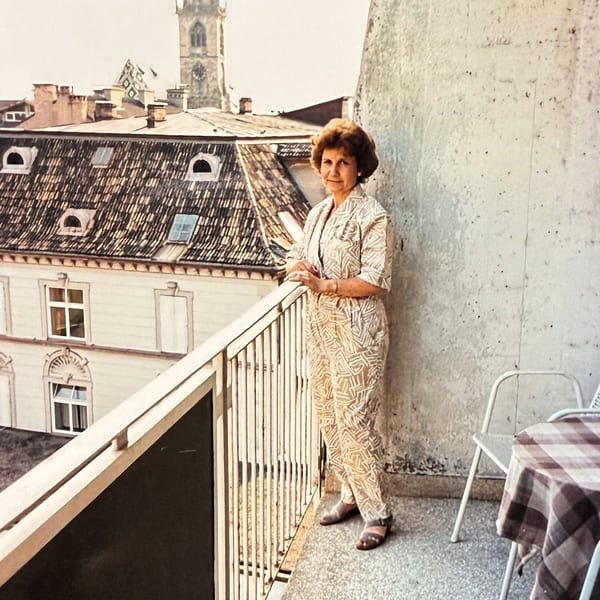 A woman in a patterned jumpsuit stands on a balcony overlooking a European city with a clock tower.