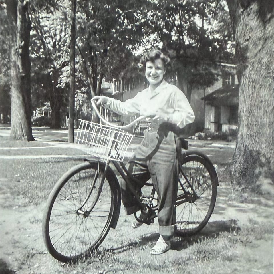 Black and white photo of a smiling woman in pants standing next to a bicycle with a basket.