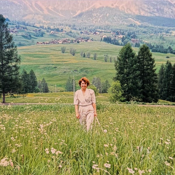 A woman in a white outfit stands in a field of tall grass and wildflowers with mountains in the background.