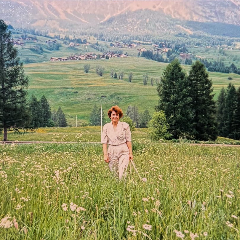 A woman in a white outfit stands in a field of tall grass and wildflowers with mountains in the background.